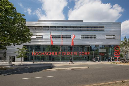 Dieses Bild zeigt das zentrale Hörsaalgebäude der Hochschule Düsseldorf mit einer Fassade aus Stahlblech und Glas. Im Erdgeschoss befinden sich die Hörsäle mit roten Stühlen. Im Obergeschoss gibt es Büroräume für die Beschäftigten der Hochschule.
